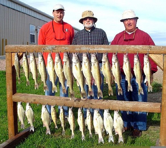 Three men stand behind a rack of caught fish. They're outdoors near a building.
