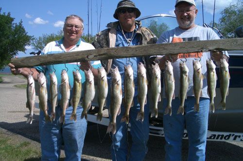 Three men display a string of caught fish, smiling near a boat and water.