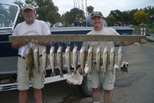 Two men holding a wooden board with numerous fish hanging from it; next to a boat at a dock.