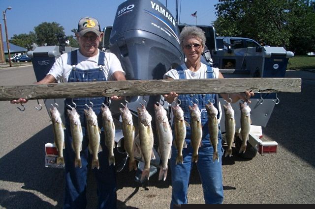 Two people holding a wooden bar with several fish hanging from it; boat in background.