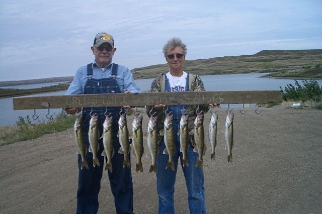 Two people hold up a board with several fish hanging from it; outdoor setting with water and sky.