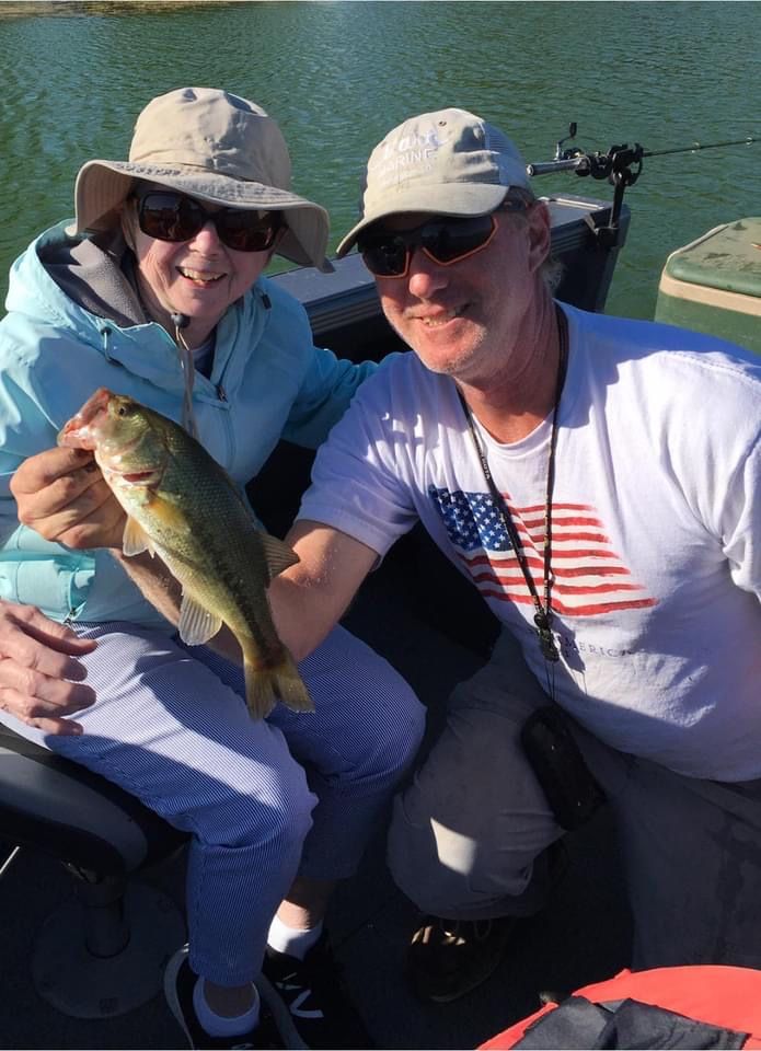 Couple in boat, holding up a fish. Man in white shirt and sunglasses, woman in hat and blue jacket.