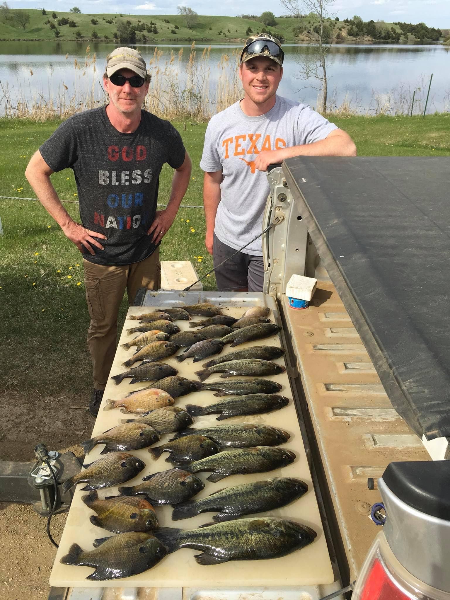Two men pose with a large catch of fish on a tailgate beside a lake.