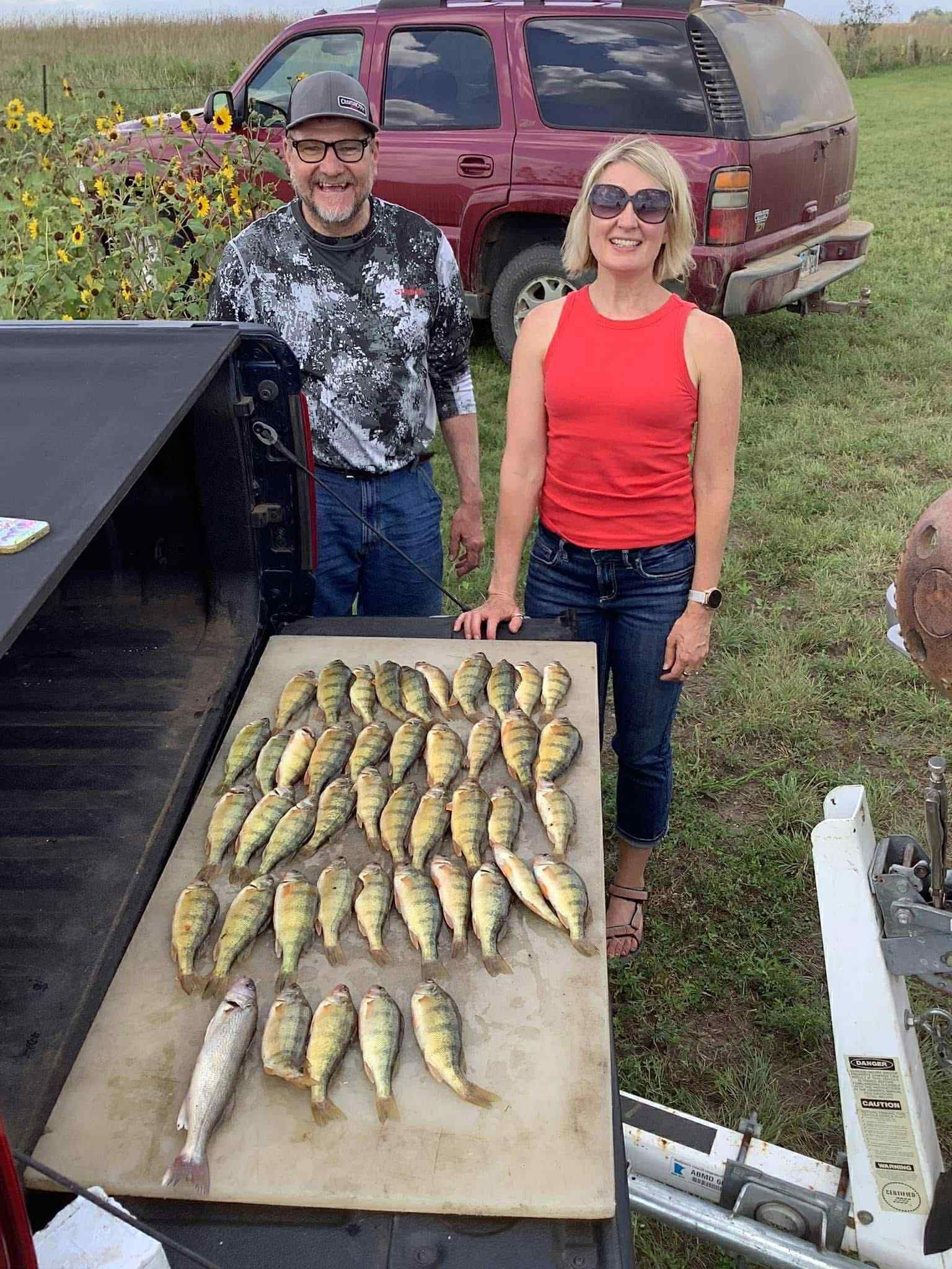 Man and woman with many yellow perch on a cutting board, truck in background.
