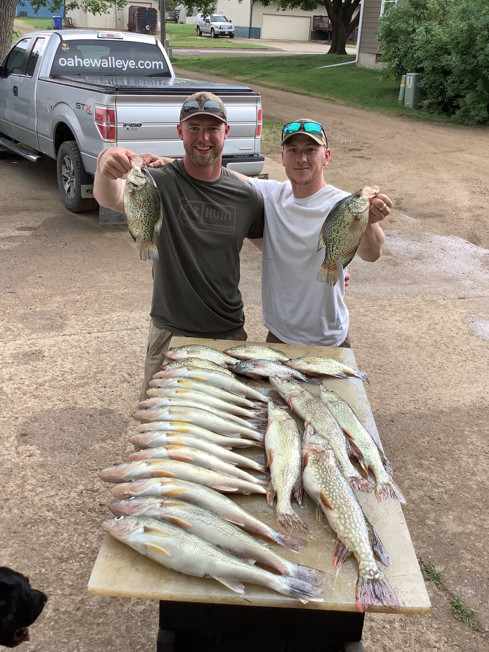 Two men display a large catch of fish on a table outside; a Ford F-150 truck is parked in the background.