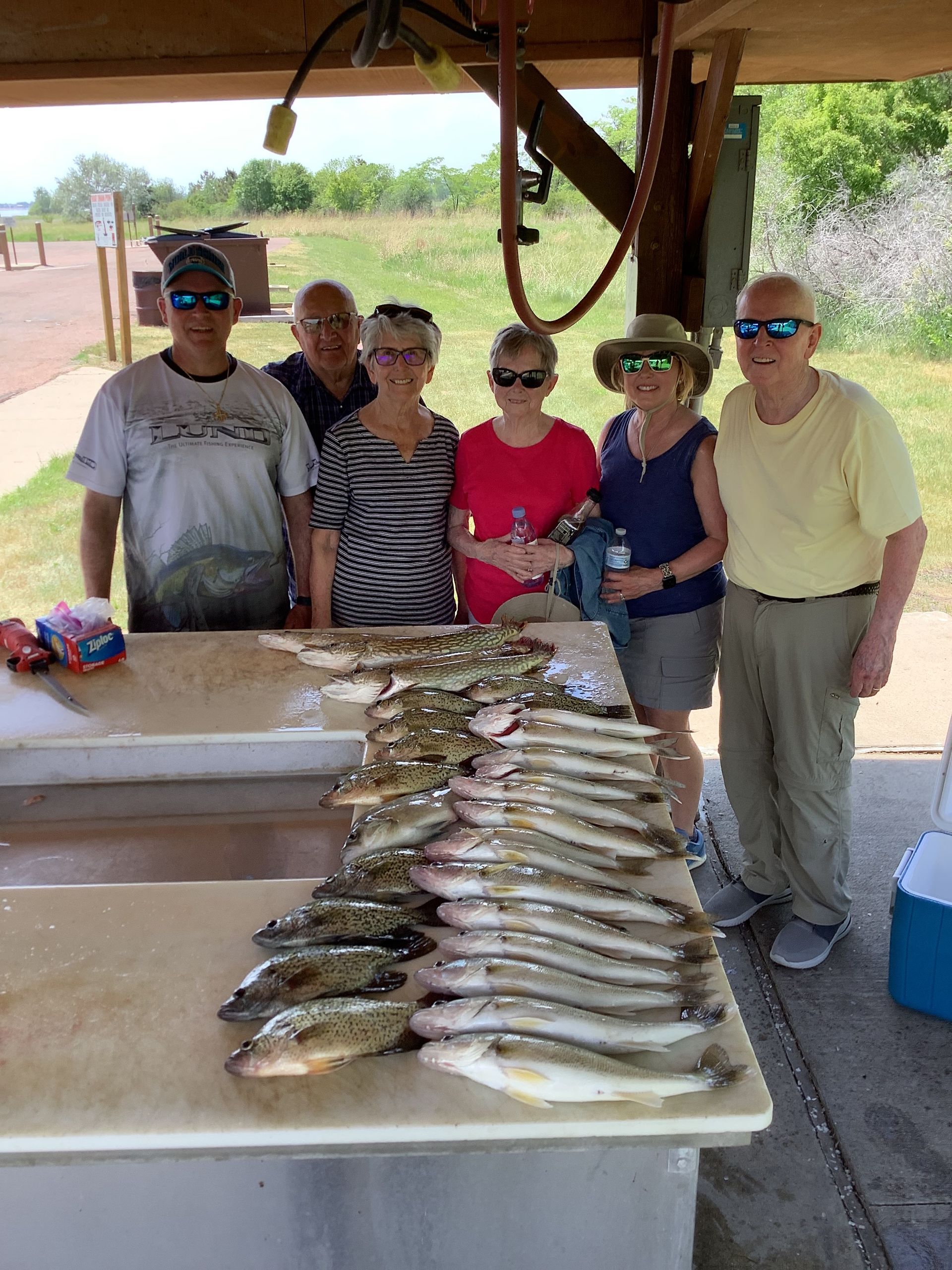 Group of people pose with a large catch of fish on a cleaning table outdoors.