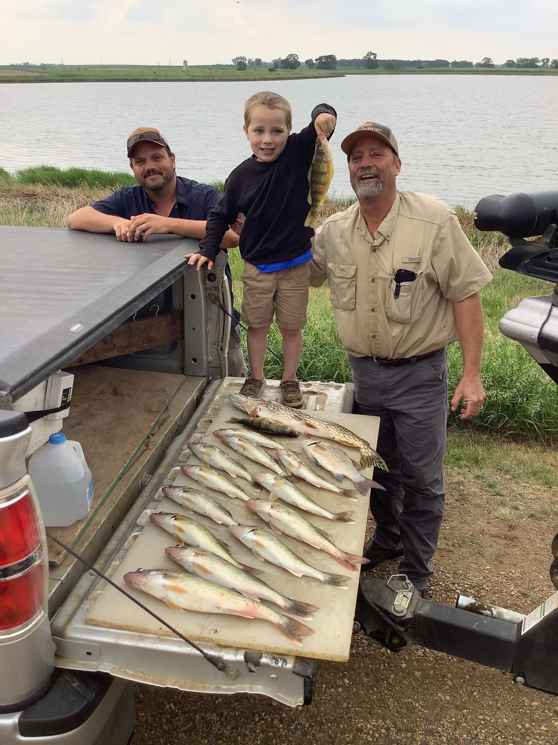 Three people pose with a catch of fish next to a truck. A boy holds up a fish, smiling.