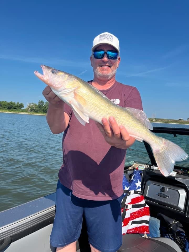 Man on a boat holds up a walleye fish against a blue sky.