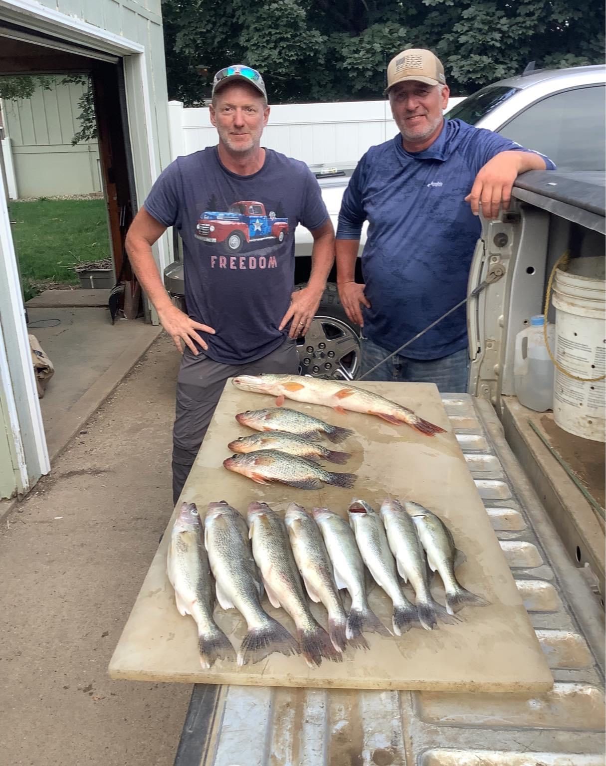 Two men stand behind a truck bed with a cutting board displaying freshly caught fish.