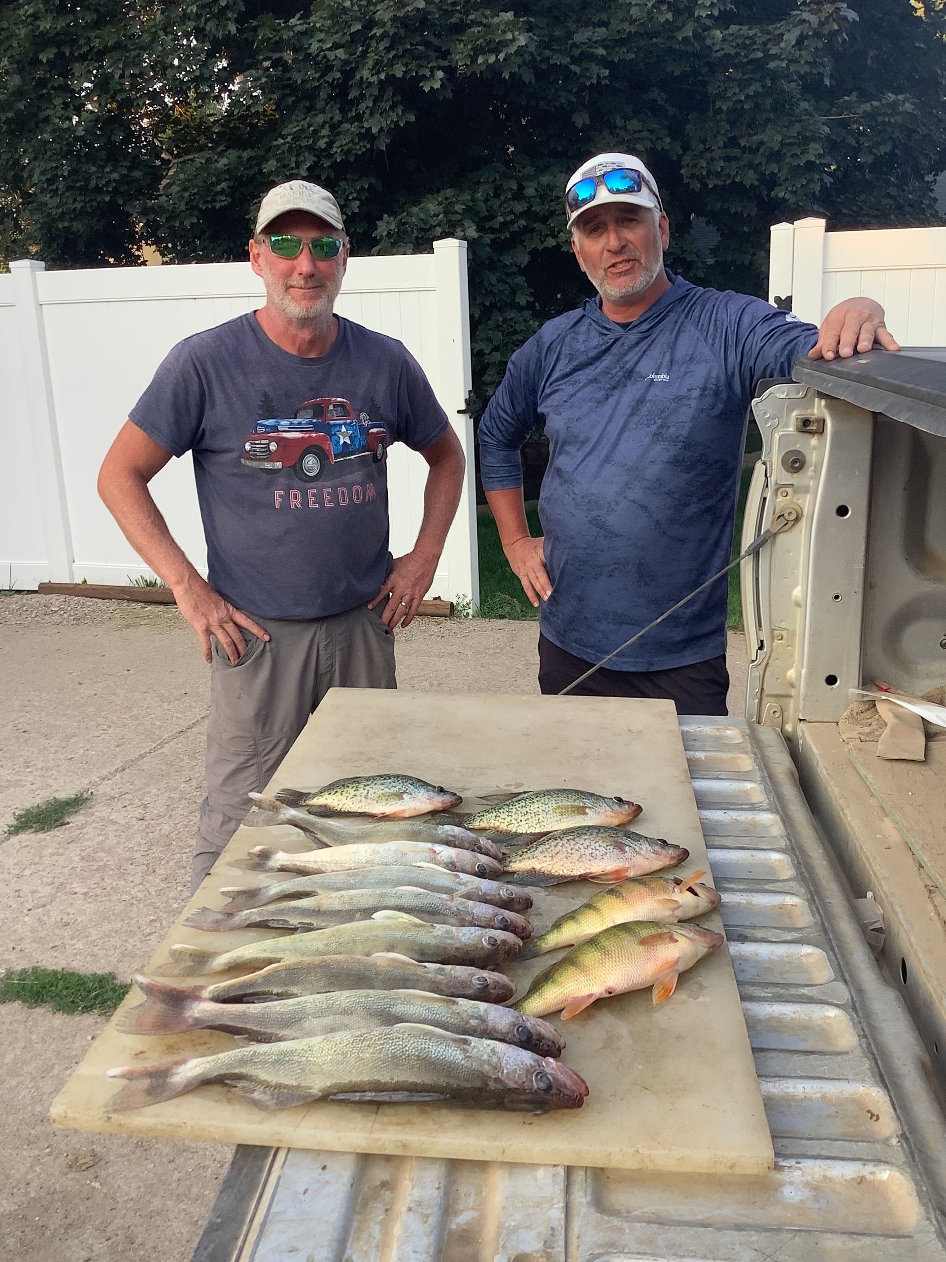 Two men stand beside a RAM truck bed displaying freshly caught fish on a cutting board, outdoors.
