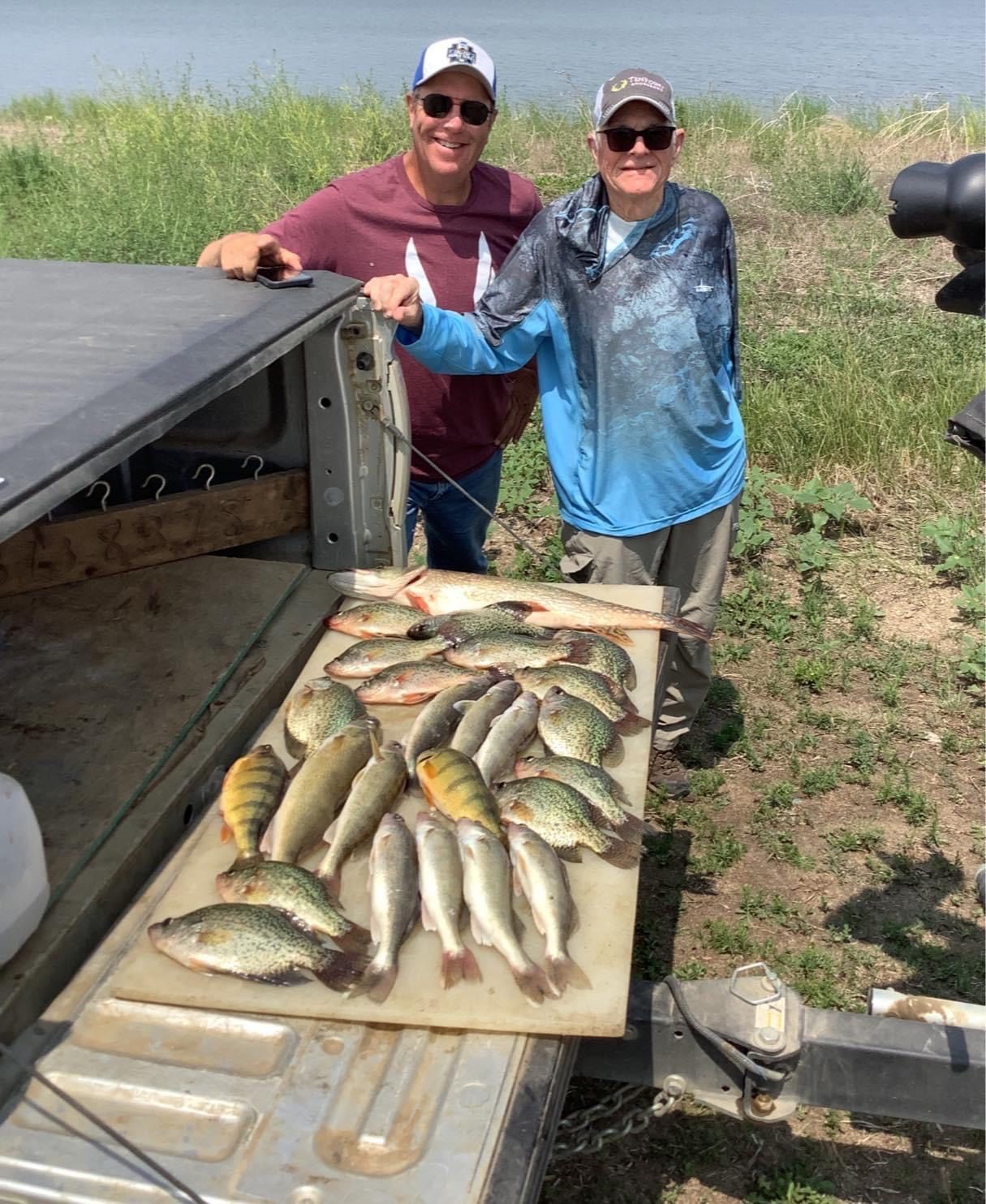 Two men beside a truck bed display a large catch of fish near a body of water.