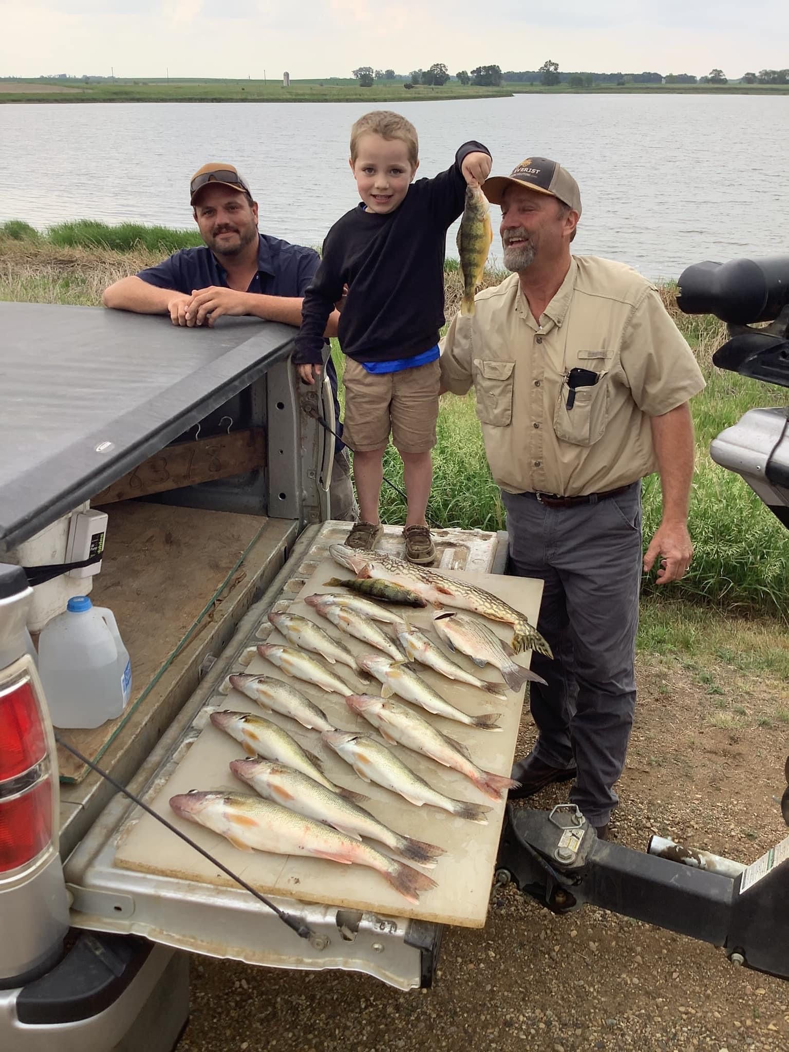 Three people, including a child, pose with a catch of fish on a truck tailgate by a lake.