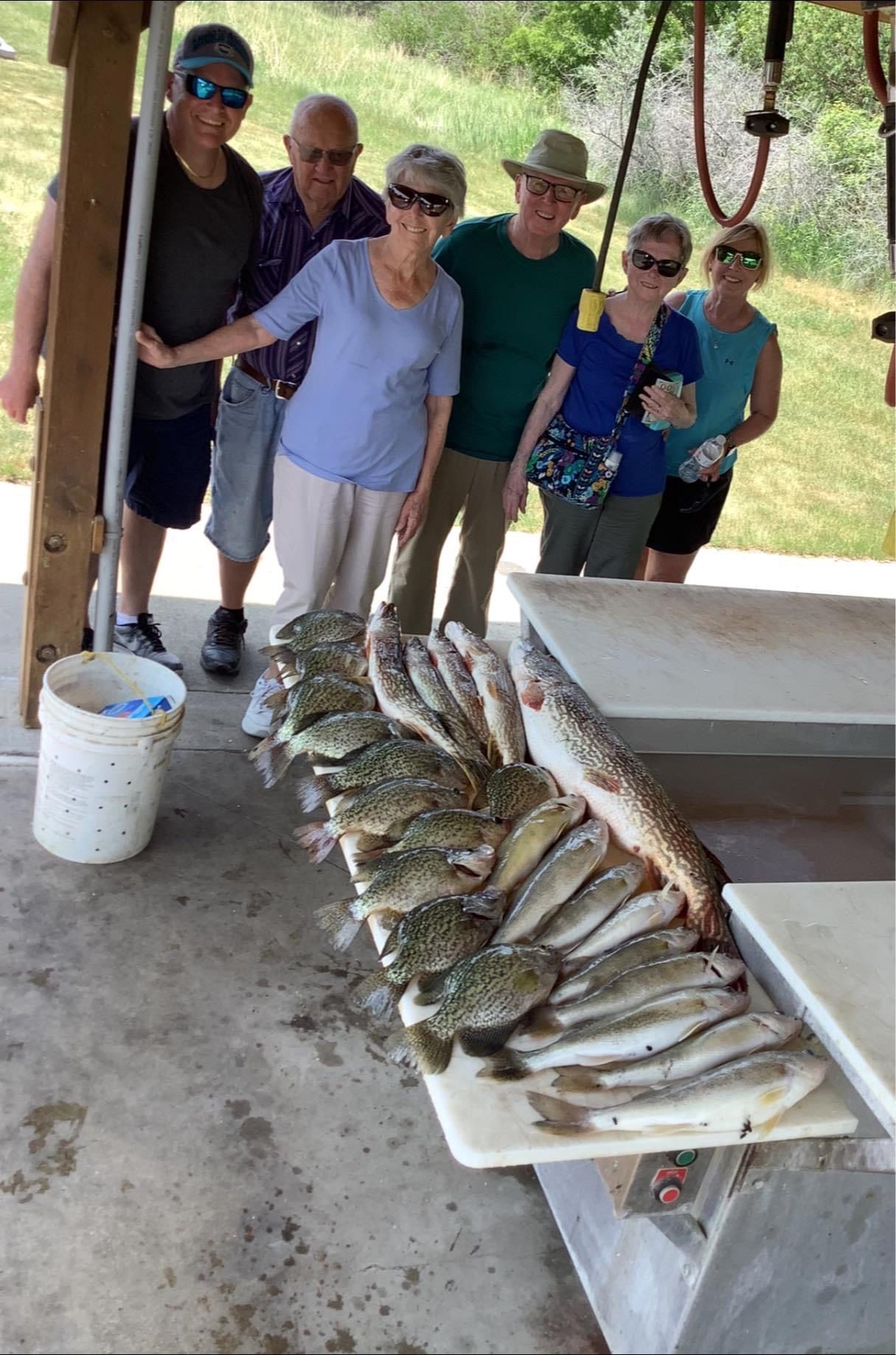 Group poses with caught fish on a cleaning table outdoors. Several people smile.