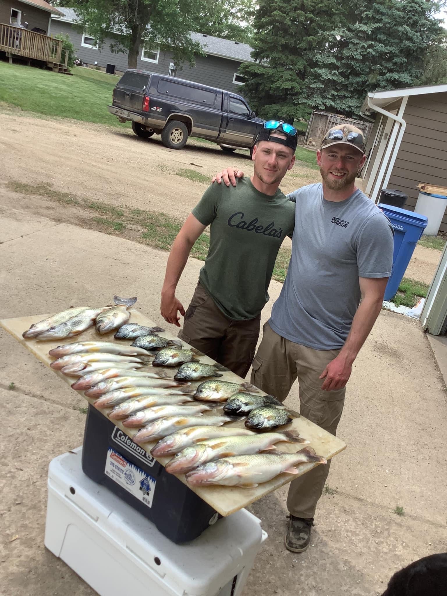 Two men pose with a large fish catch laid out on a table, outdoors near a truck and houses.