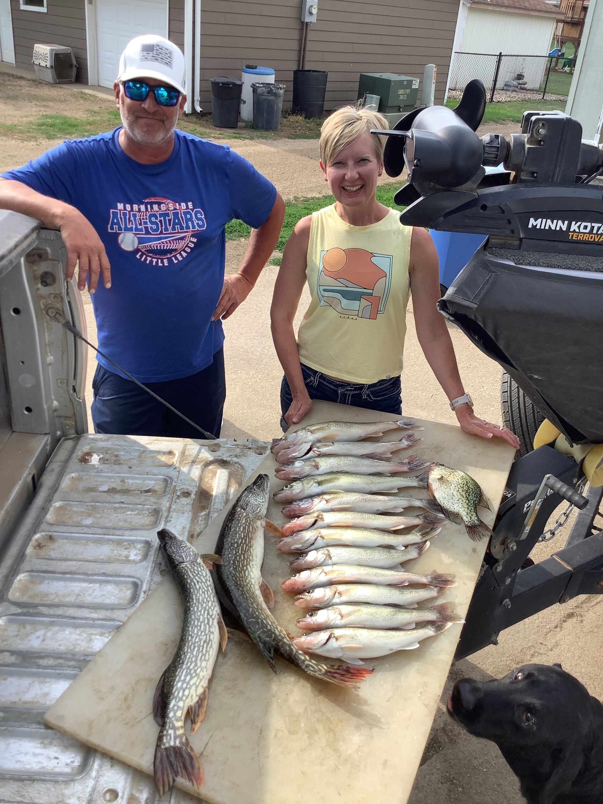 Two people stand by a truck bed and MinnKota trolling motor with a large catch of fish, smiling. Sunny outdoor setting.