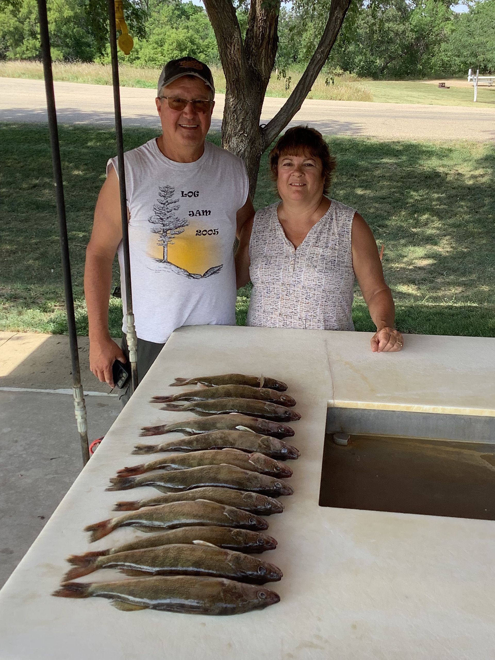 Couple poses with a row of fish on a white table under a tree, smiling outdoors.