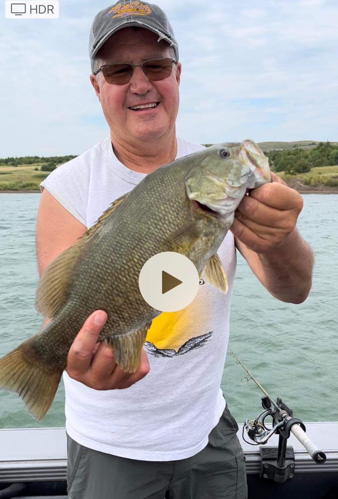Man holding a fish on a boat. The fish is greenish brown. He is smiling. Outdoors setting.