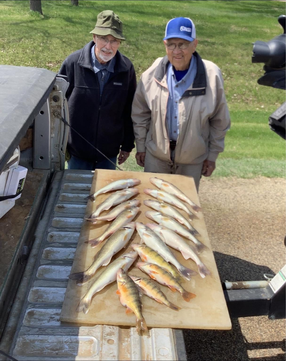Two men stand behind a truck bed with a cutting board displaying freshly caught fish.