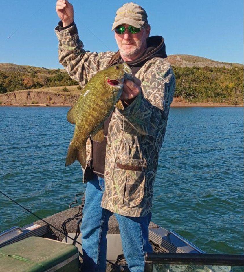 Man in camo jacket on a boat holding a largemouth bass, raising arm in a gesture of success, on a lake.