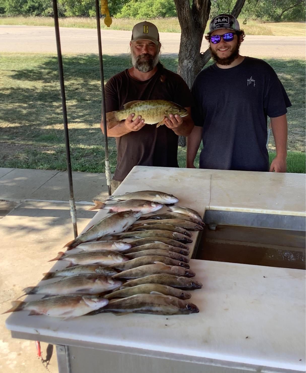 Two men display fish catch. One holds large fish. Many other fish lay on a table outside.