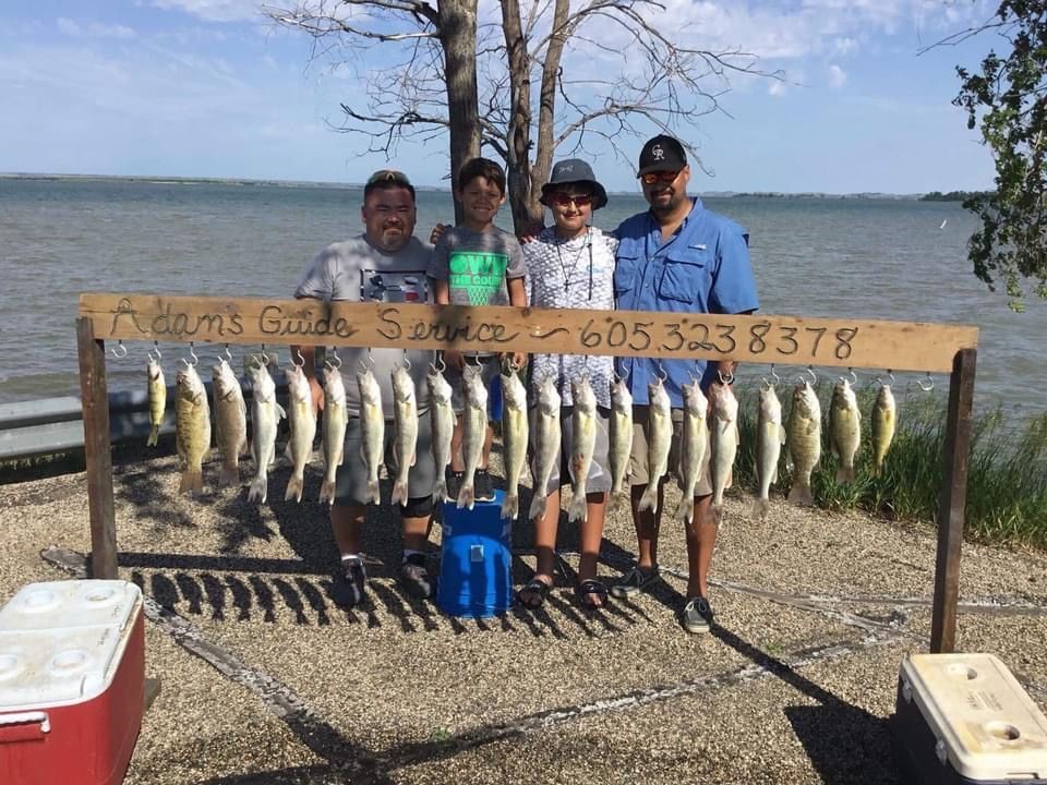 Four people posing with a rack of caught fish by a lake. A sign advertises fishing guide service.