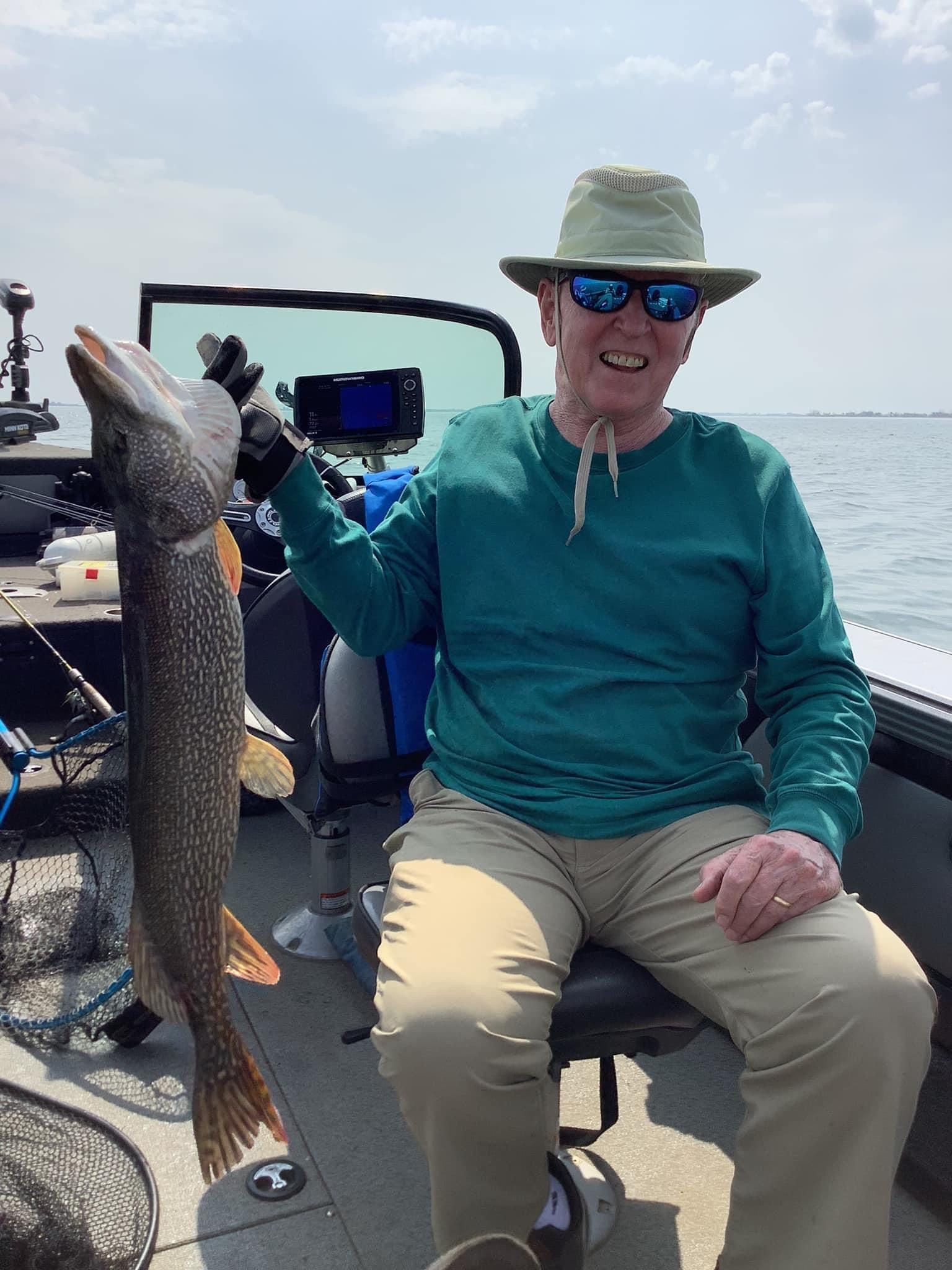 Man on a boat holds up a large fish, smiling. He wears sunglasses, a hat, and a teal shirt.