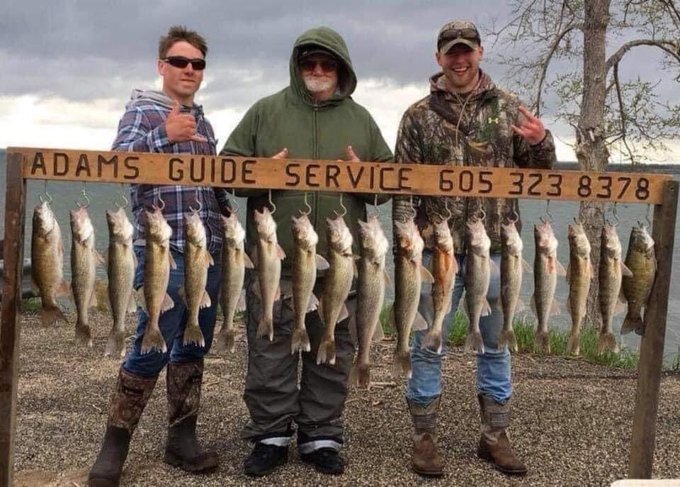Three men with fishing catch, displayed on a rack, near water. “Adams Guide Service” sign with phone number.