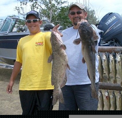 Two men holding up catfish they caught, near a boat, sunny day.