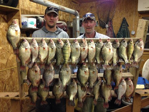 Two men display a large catch of fish on a wooden rack inside a cabin.