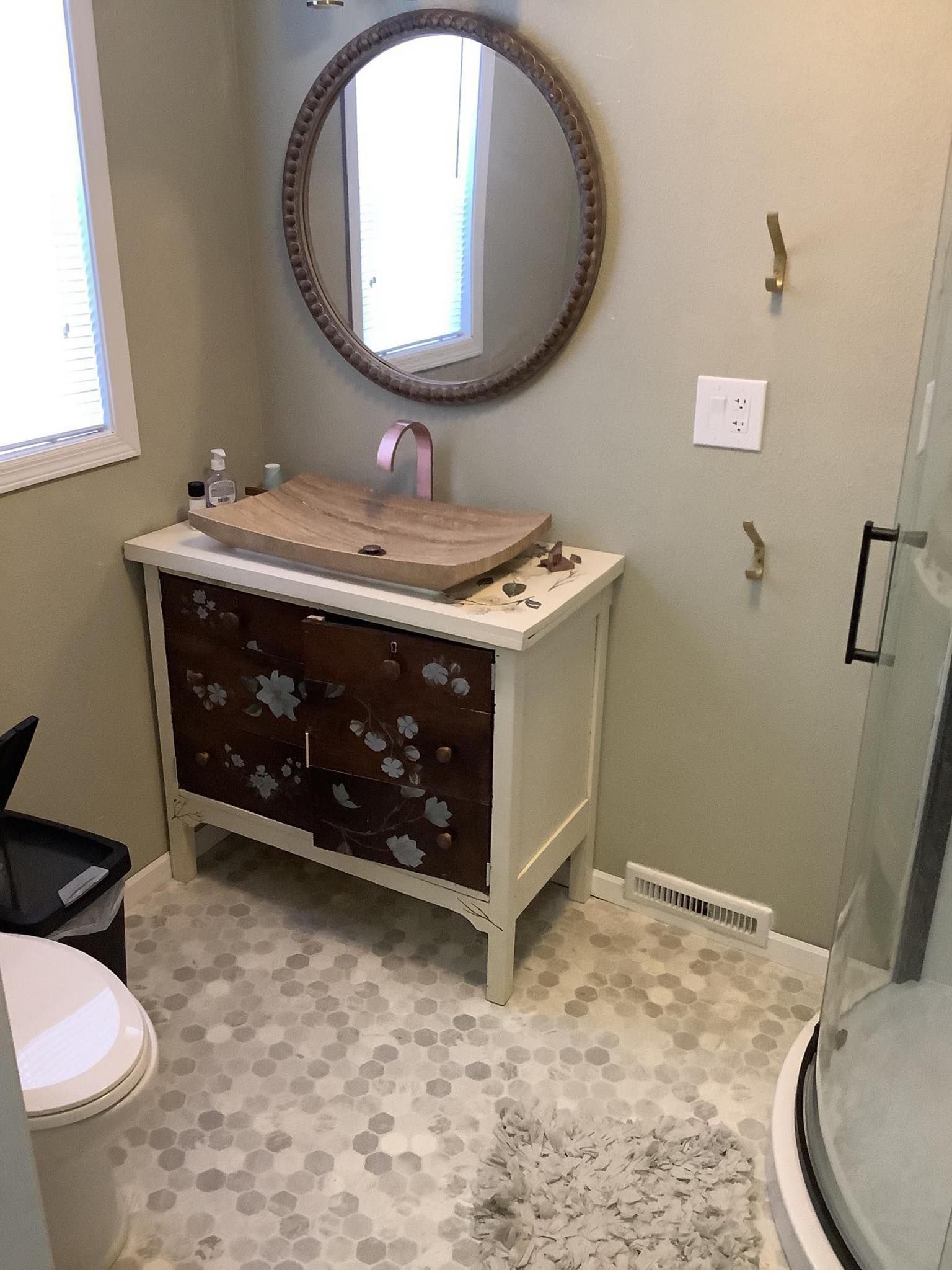 Bathroom with wooden vanity, round mirror, floral accents, hexagonal tile flooring, and a shower.