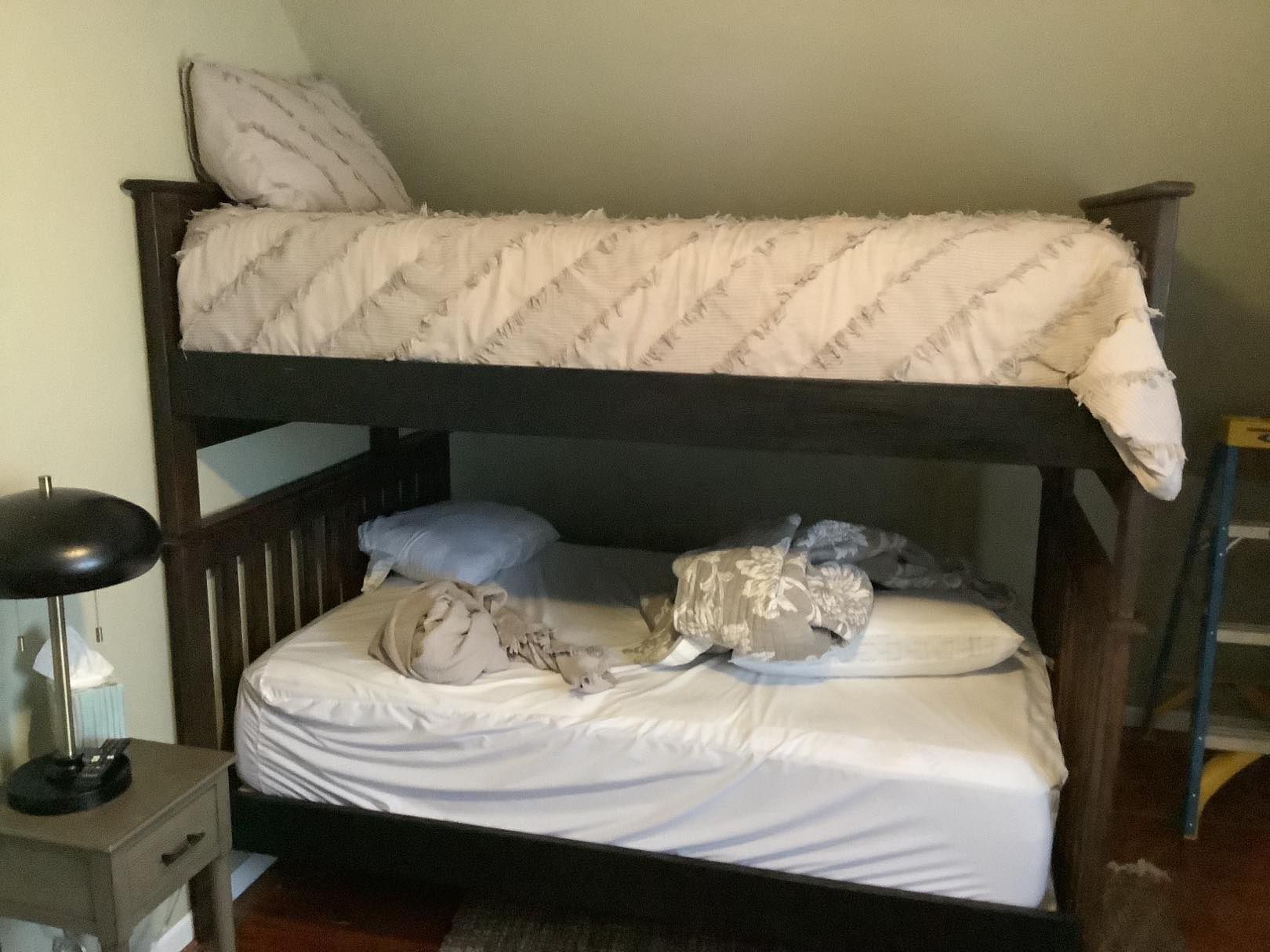 Bunk bed with a dark frame and white bedding, in a room with a nightstand and lamp.