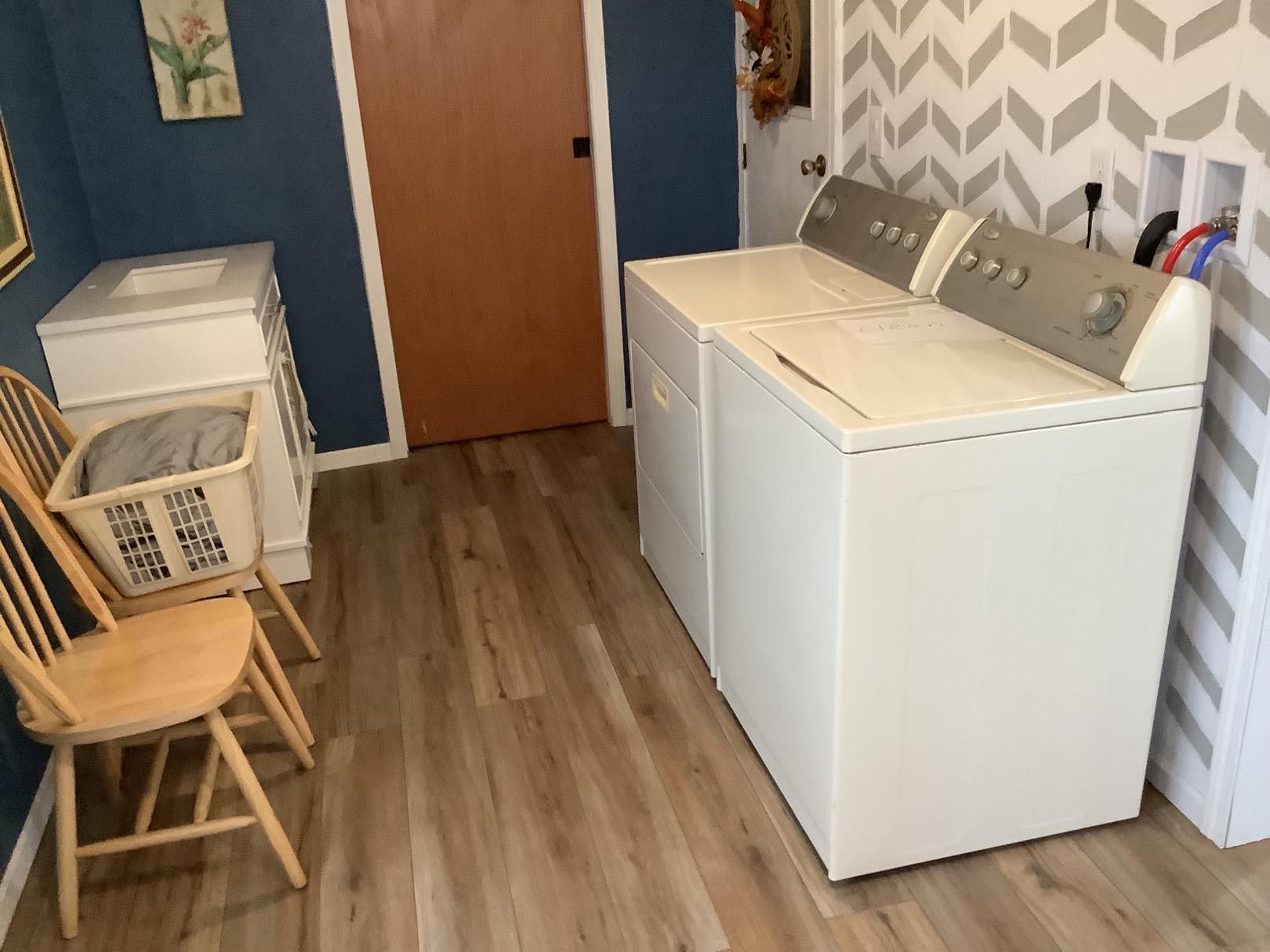Laundry room with a washer, dryer, wooden chair, and a cabinet. Chevron patterned wallpaper and wooden floors.