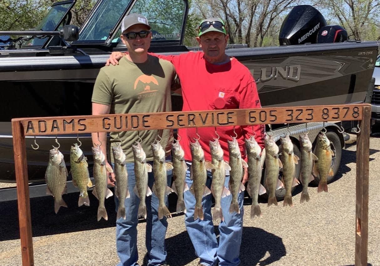 Two men pose with caught fish; a sign says Adams Guide Service, boat in background.