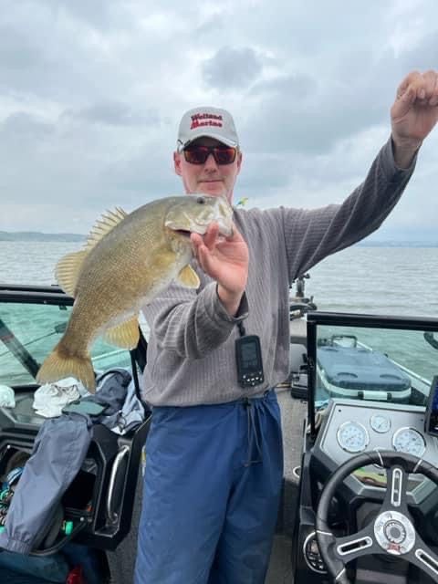 Man holding a smallmouth bass on a boat, overcast sky, lake in the background.