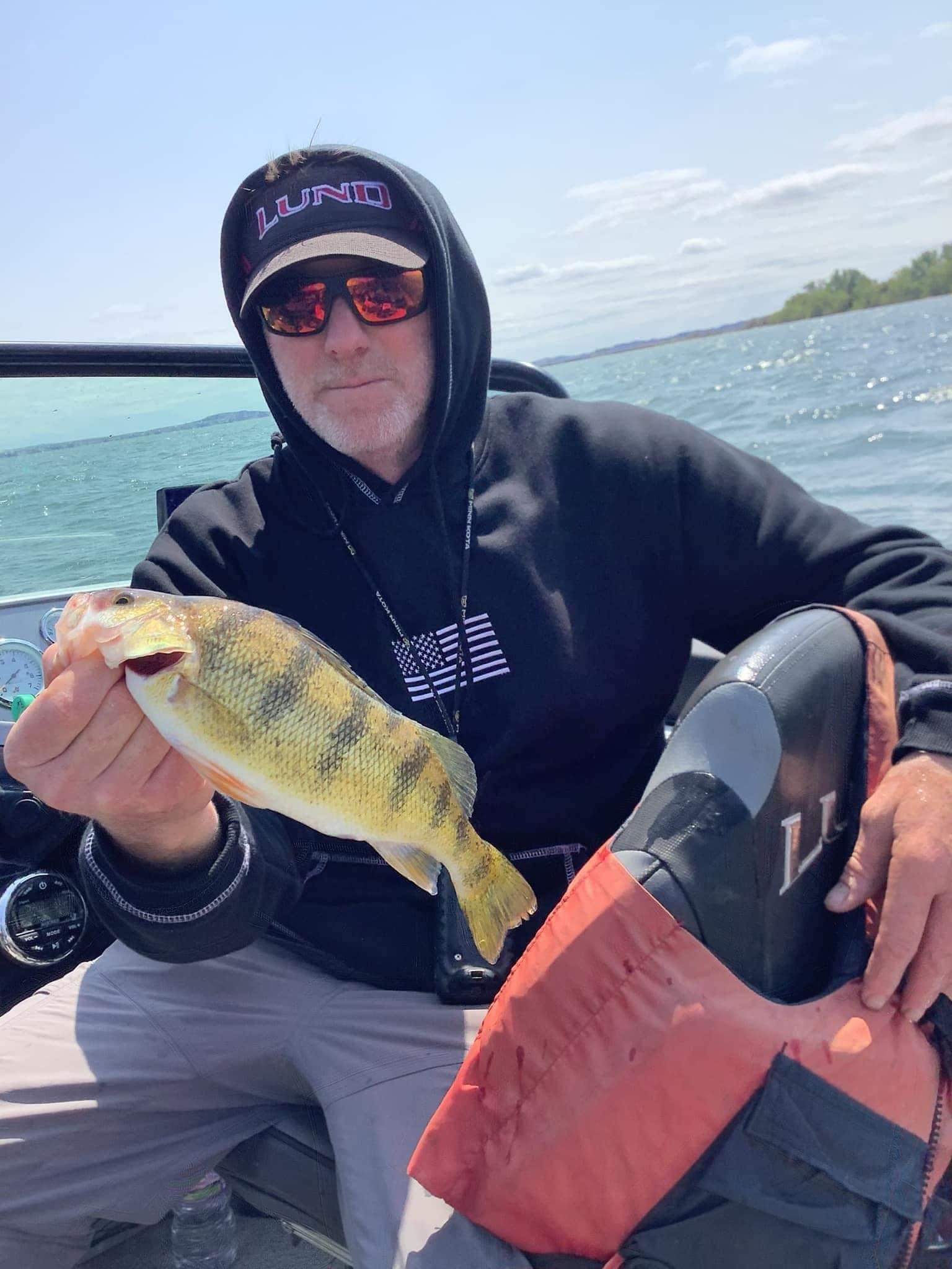 Man in a boat holding a yellow perch. He wears a black hoodie, sunglasses, and a hat; water and blue sky in the background.