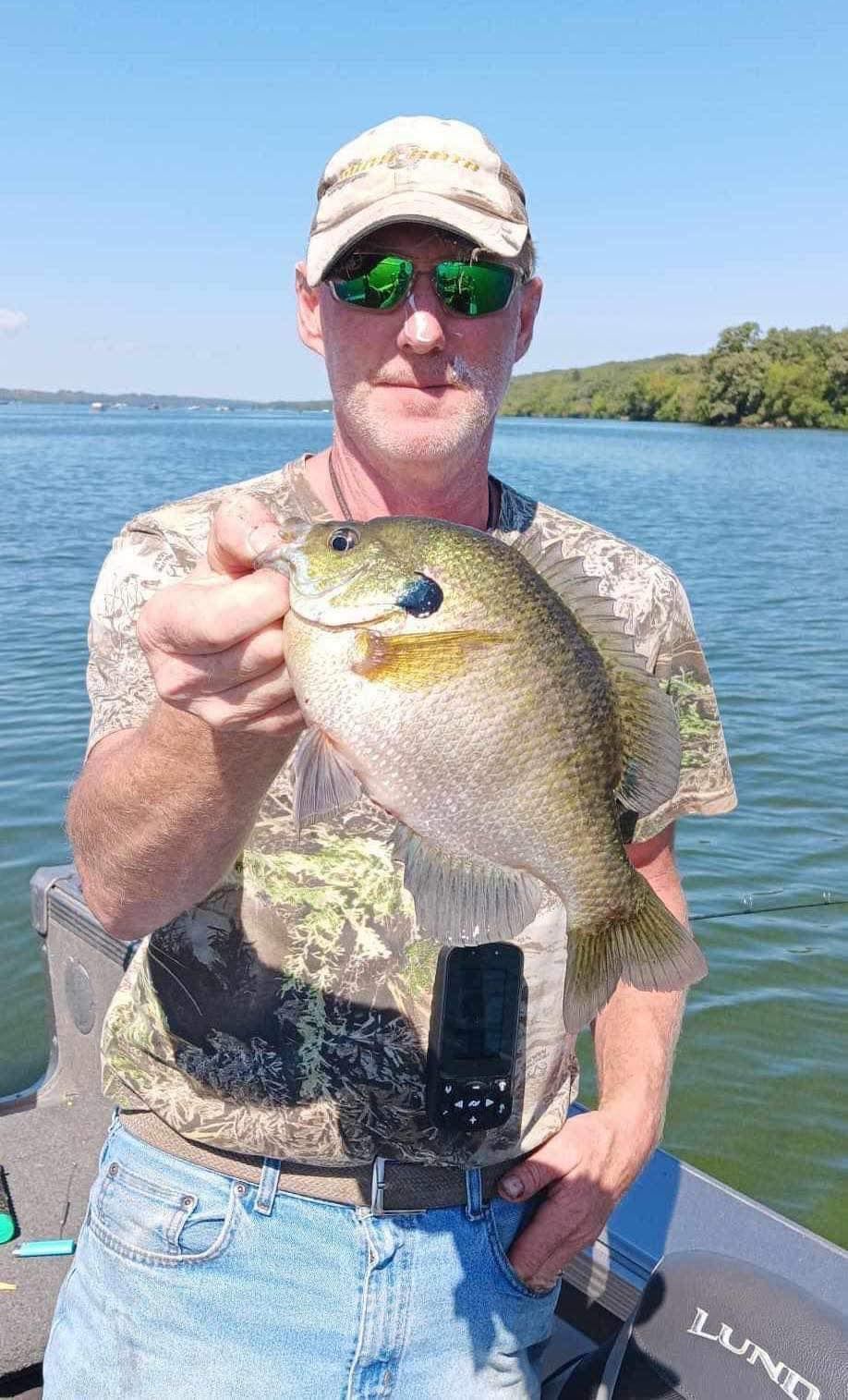 Man in sunglasses holding a large bluegill fish on a lake. He's wearing a camouflage shirt and hat.