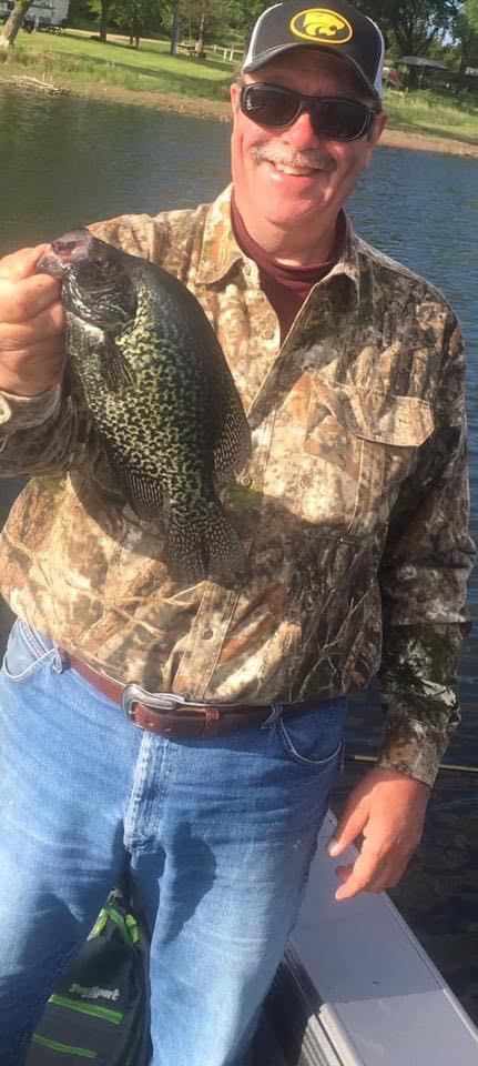 Man holding a speckled fish, smiling. He wears sunglasses, a cap, camouflage shirt, and jeans. Outdoors, water visible.