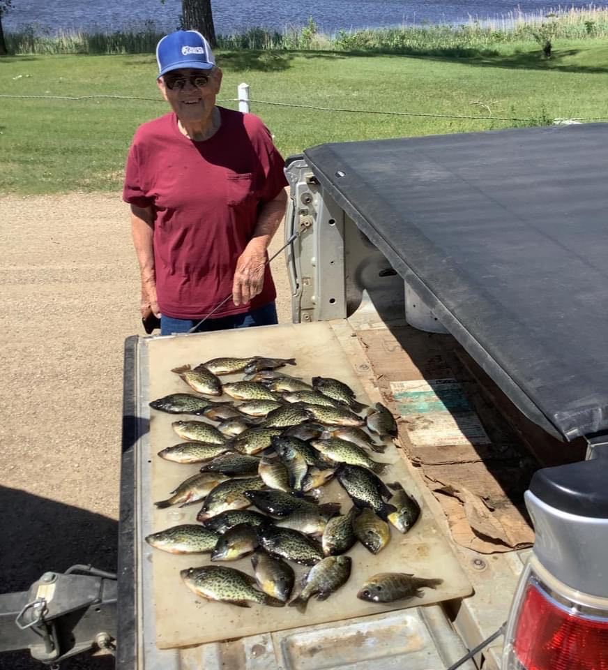 Man in a cap stands by a truck bed filled with freshly caught fish on a sunny day near water.