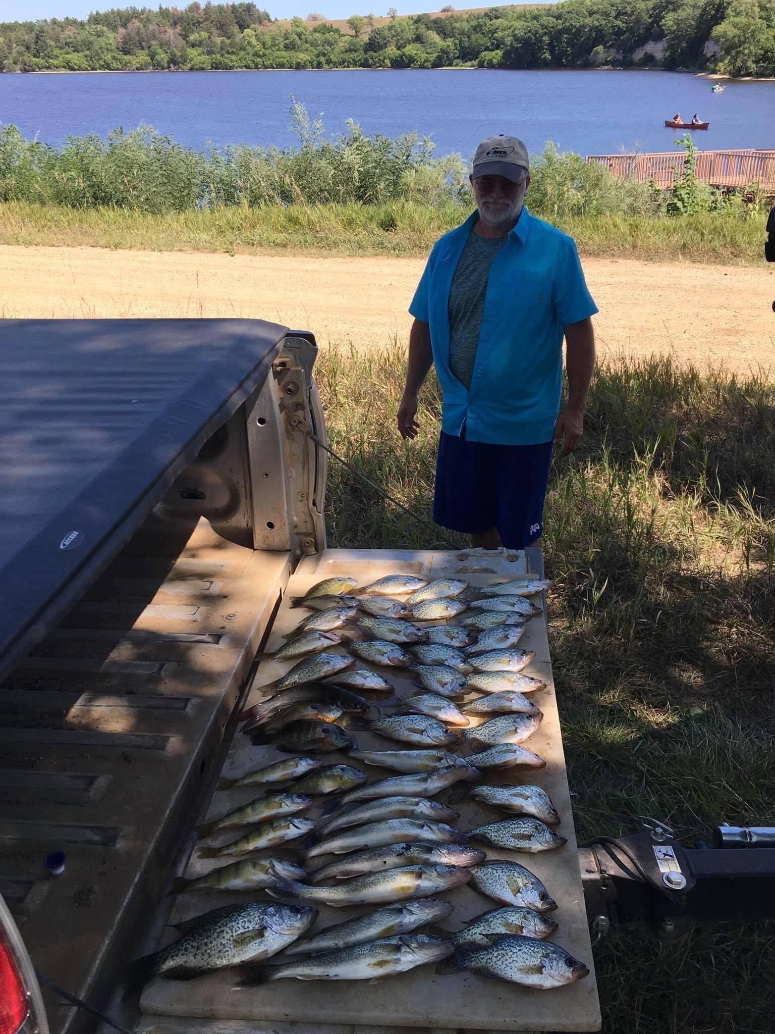 Man stands near truck bed with a large pile of caught fish, overlooking a lake.