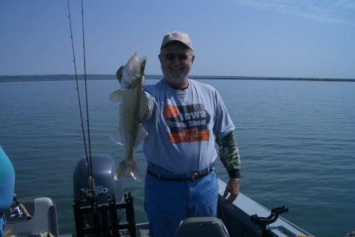 Man on a boat holding a large fish, posing against a body of water on a sunny day.