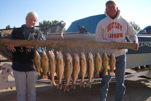 Two people hold a stringer with many caught fish, boat in background. Sunny day.
