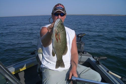 Man on a boat holding up a freshly caught South Dakota bass with a calm lake in the background.