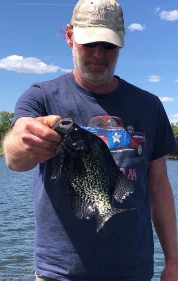 Man in blue shirt holding a black crappie fish, on a sunny day.