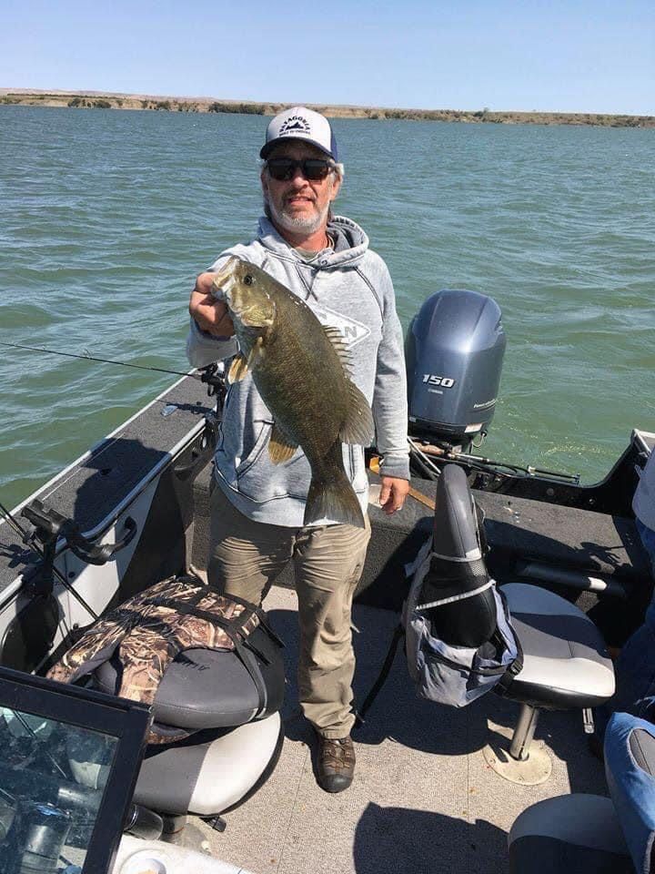 Man holding a fish on a boat. The water is visible in the background. Sunny day.