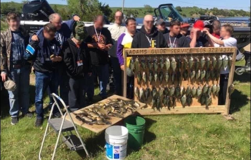 Group of people posing with a wooden display of fish after fishing. Green grass and lake in the background.