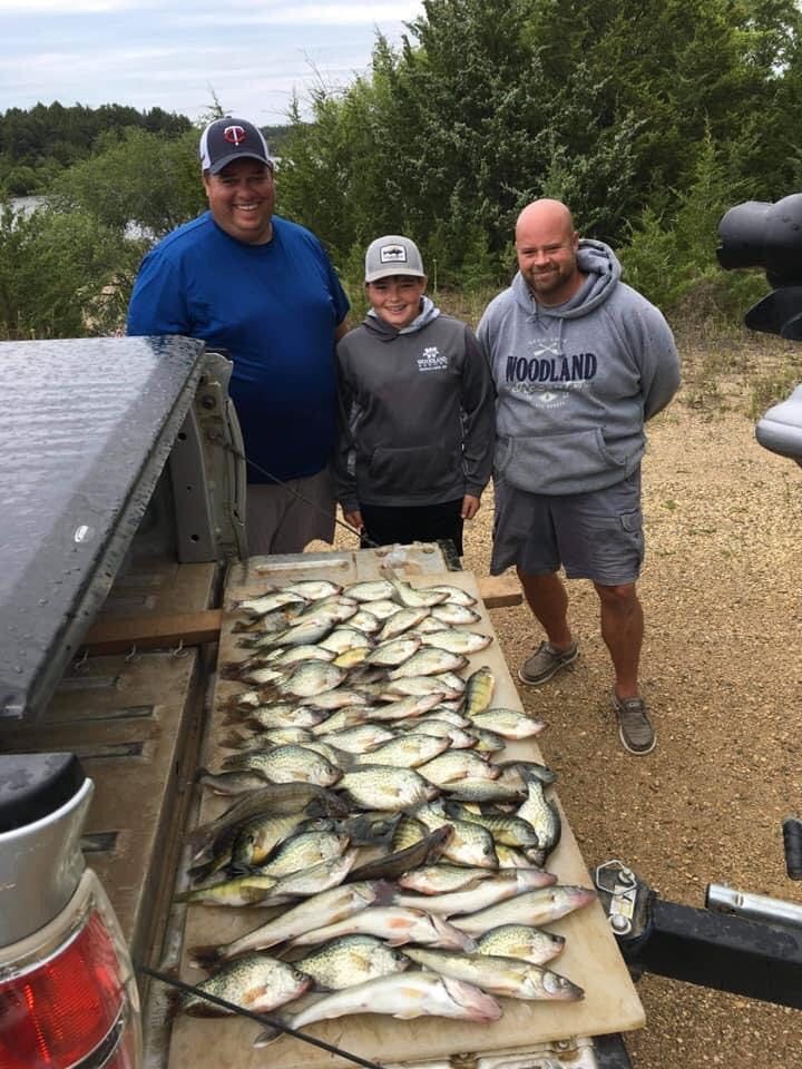 Three people pose with a large pile of fish on a surface. Two men and a youth smile outside near water and trees.