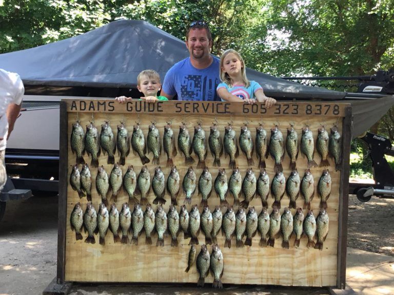 Man and two children pose with a wooden board displaying many fish. 