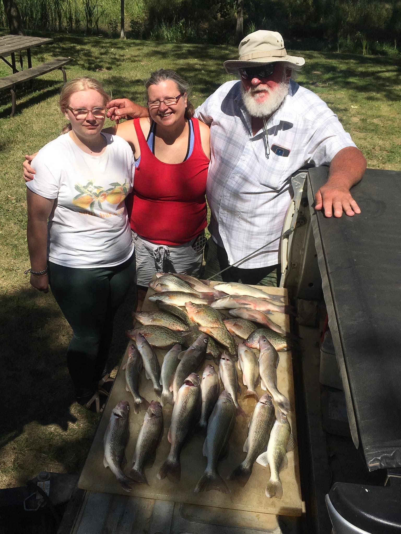 Three people pose with a large catch of fish on a wooden table outdoors.