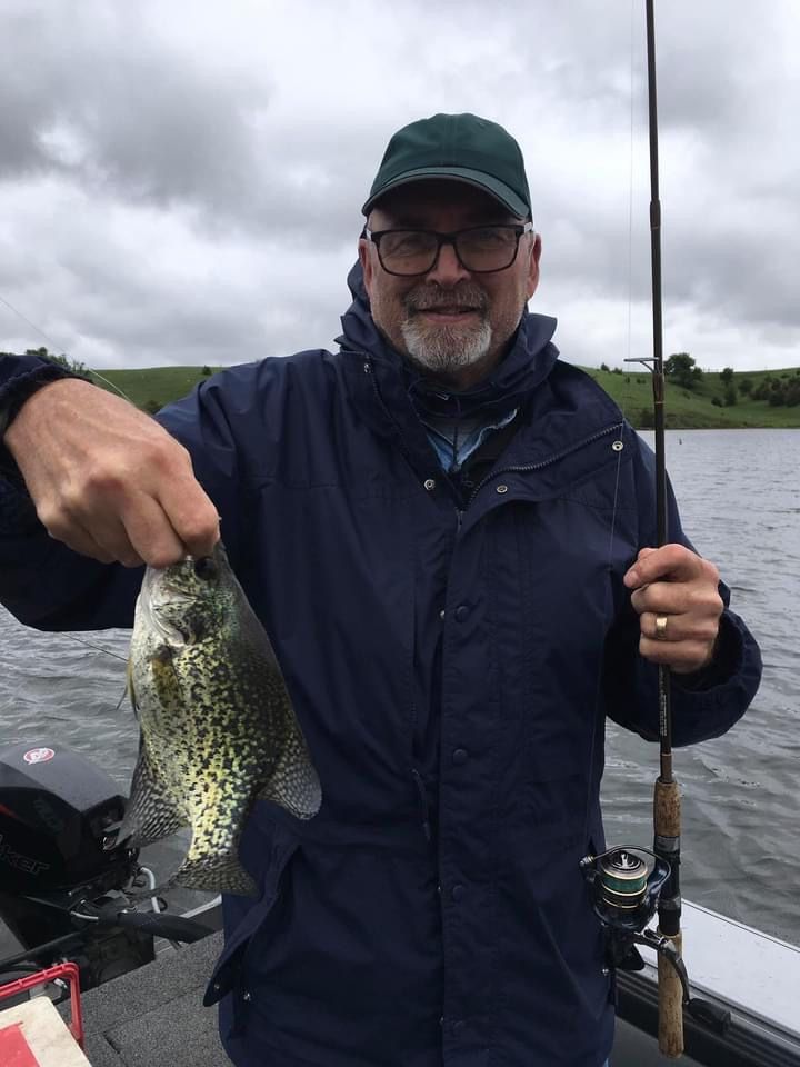Man in a boat holding a fish, cloudy day on a lake. He wears a hat and jacket, smiling.