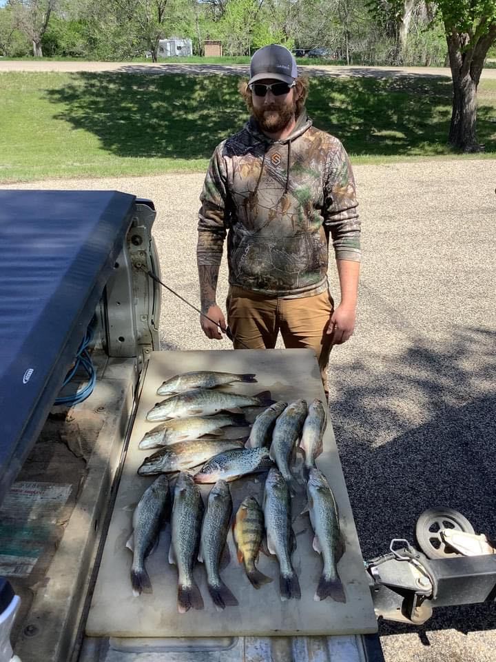 A man stands behind a truck bed with a collection of caught fish. He wears a camo hoodie.